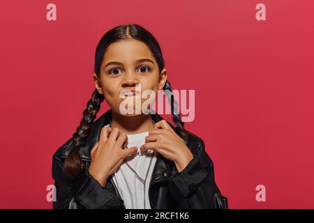 Portrait eines aggressiven und modischen jungen Mädchens mit Frisur, Lederjacke und Blick in die Kamera, während sie isoliert auf Rot, Mädchen w steht Stockfoto