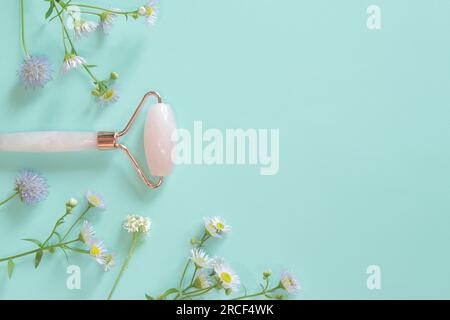 Gesichtsmassagewalze aus Rosenquarz auf blauem Hintergrund mit Blumen. Gesichtsmassagegerät Anti-Falten Anti-Aging-Hautpflegewerkzeug Stockfoto