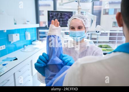 Junge Assistentin in Operationsuniform bereitet einen Chirurgen auf die Operation vor Stockfoto