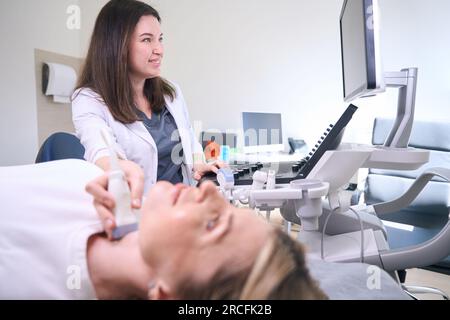 Eine angenehme Ärztin führt eine Ultraschalluntersuchung der Schilddrüse durch Stockfoto