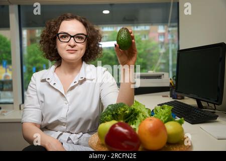 Eine lockige Frau mit Brille hält eine Avocado in der Hand Stockfoto