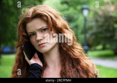 Runzelndes Mädchen mit langen Ingwerhaaren und Sommersprossen, das im Sommerpark mit dem Handy spricht und in die Kamera schaut Stockfoto