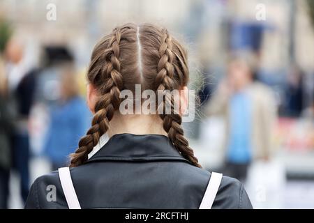 Ein Mädchen mit geflochtenen Zöpfen, das eine Lederjacke auf der Straße trägt. Weibliche Frisur und Mode in der Stadt Stockfoto