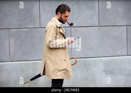 Ein Mann mit einer rauchenden Pfeife, der einen Regenmantel trägt und auf den Smartphone-Bildschirm schaut, während er auf einer Straße steht Stockfoto