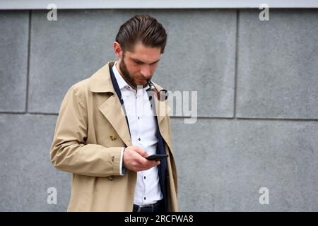 Ein Mann mit einer rauchenden Pfeife, der einen Regenmantel trägt und auf den Smartphone-Bildschirm schaut, während er auf einer Straße steht Stockfoto