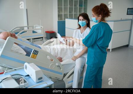 Ärztinnen mit einem Blatt Papier, die mit dem männlichen Patienten in der Kammer sprechen Stockfoto