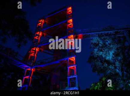 Beelitz, Deutschland. 14. Juli 2023. Der Aussichtsturm auf dem Gelände des Baumwipfels „Baum & Zeit“ in Beelitz-Heilstätten ist abends rot beleuchtet. An drei Abenden im Juli und August bietet der Abenteuerbereich seinen Gästen ein Abendprogramm mit Musik, Lichtinstallationen und Vorstellungen. Kredit: Monika Skolimowska/dpa/ZB/dpa/Alamy Live News Stockfoto