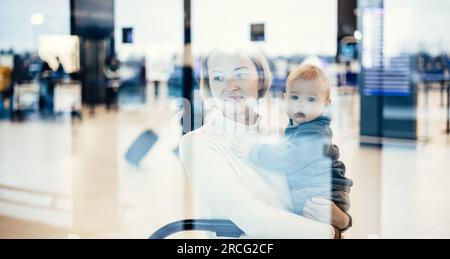 Die nachdenkliche junge Mutter schaute durch das Fenster und hielt sein kleines Kind fest, während sie darauf wartete, an den Abflugsteigen des Flughafenterminals an Bord eines Flugzeugs zu gehen. Das „Reise mit Baby“-Konzept Stockfoto