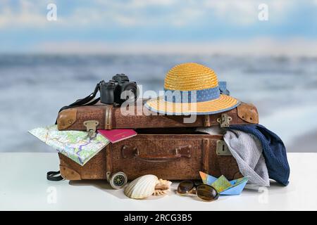 Oldtimer-Koffer mit Reiseaccessoires für den Strandurlaub wie Strohhut, Fotokamera, Handtücher, Reisepass, Karte und Sonnenbrille, Verschwommenes Meer Stockfoto