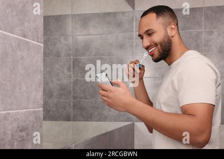 Glücklicher Mann, der sein Smartphone benutzt, während er im Badezimmer Zähne putzt, Platz für Text. Internetsucht Stockfoto