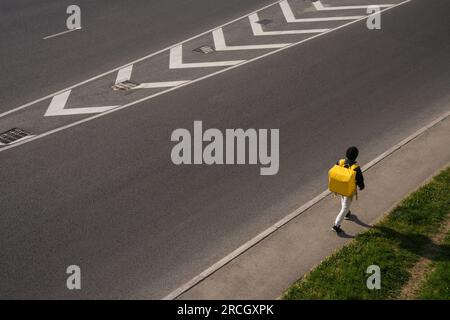 Ein Mann geht mit einer gelben Liefertasche auf dem Bürgersteig an der Straße entlang. Stockfoto