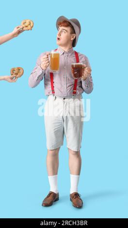 Junger Mann in traditioneller deutscher Kleidung mit Bier, der Brezeln auf blauem Hintergrund bekommt Stockfoto