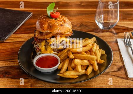 Nahaufnahme eines hausgemachten, frischen Burgers mit Rindfleisch, Zwiebeln, Tomaten, Salat, Käse und Tomatensoße. Serviert mit Kartoffelpommes und Pommes frites auf Holztabellen Stockfoto