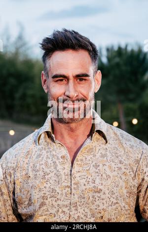 Vertikales Einzelbild eines gutaussehenden Mannes mittleren Alters, der draußen steht, lächelt und sorglos und positiv in die Kamera schaut. Positiver, glücklicher, selbstbewusster Mann mit Smiley-Einstellung. Hochwertiges Foto Stockfoto