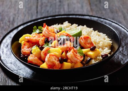 Karibische gegrillte Garnelen mit schwarzen Bohnen, Mango-Ananassalsa serviert mit braunem Reis in schwarzer Schüssel auf dunklem Holztisch, Landschaftsblick Stockfoto