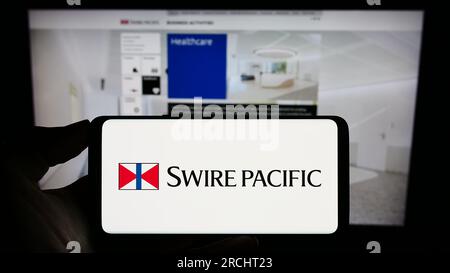 Person, die ein Mobiltelefon mit dem Logo des asiatischen Unternehmens Swire Pacific Limited auf dem Bildschirm vor der geschäftlichen Webseite hält. Konzentrieren Sie sich auf das Display des Telefons. Stockfoto