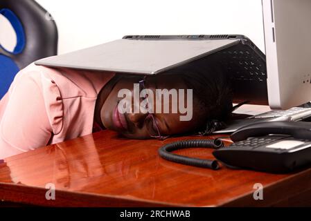 Porträt einer jungen, schönen, müden Geschäftsfrau, die am Schreibtisch sitzt und mit einem Laptop auf dem Kopf schläft. Stockfoto