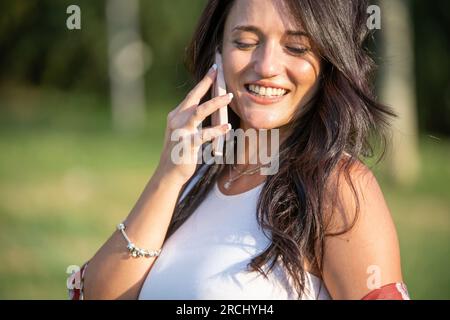 Nahaufnahme einer Frau, die lacht, während sie draußen in einem Park telefoniert. Technologiekonzept. Stockfoto