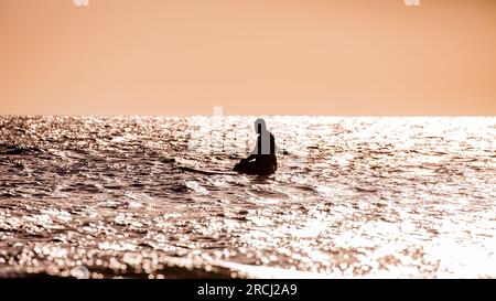 Silhouette einer Person, die auf einem Surfbrett auf dem Ozean saß. Der Himmel ist klar und wartet auf eine Welle Stockfoto