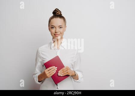 Selbstbewusste Buchhalterin in weißem Hemd, Haare in Brötchen, hält rotes Notizbuch mit Firmenunterlagen und Transaktionen, schaut in die Kamera, isoliert auf grauem Rücken Stockfoto