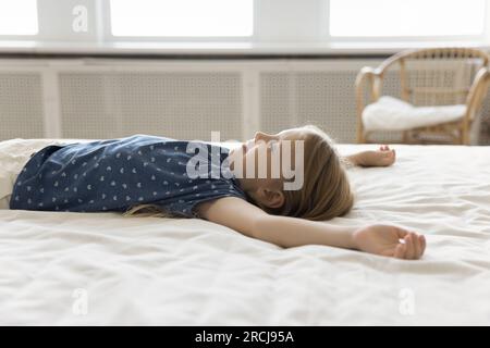 Positives kleines Mädchen, das sich auf dem Bett mit weißer Bettwäsche ruht Stockfoto