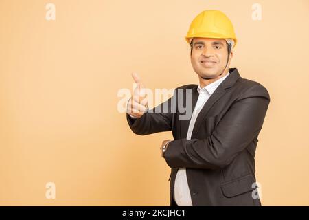 Glücklicher indischer Ingenieur mit schwarzem Anzug und gelbem Schutzhelm, der die Daumen hoch und isoliert auf beigefarbenem Hintergrund macht. Speicherplatz kopieren. Stockfoto