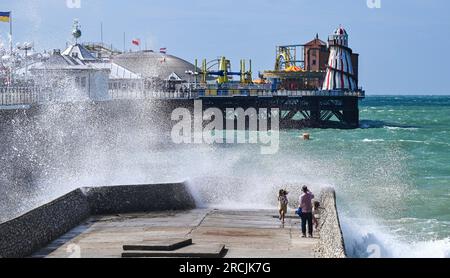 Brighton UK 15. Juli 2023 - Besucher werden nass, wenn sie sehen, wie riesige Wellen am Brighton Beach und am Ufer des Piers krachen, als ungewöhnlich starker Wind für Juli Batter Teile des Vereinigten Königreichs : Credit Simon Dack / Alamy Live News Stockfoto