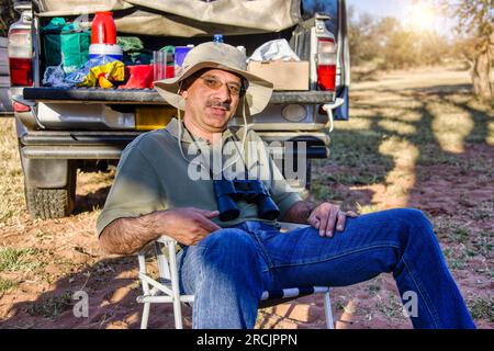 ein arabischer Mann mit Fernglas und Safarihut saß auf einem Stuhl vor seinem beladenen Geländewagen und machte eine Grillpause Stockfoto