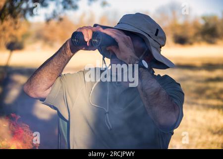 ein arabischer Mann mit Fernglas und Safarihut neben dem Lagerfeuer, der die Vögel beobachtet Stockfoto