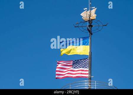 Die Flaggen der Ukraine und der USA flattern auf einem Fahnenmast vor einem blauen Himmel. Statussymbol. Ukrainische blaue und gelbe Flagge. Windtag im Freien. Stockfoto