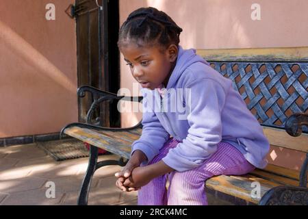 Kleines afrikanisches Mädchen mit Zöpfen, das auf der Bank vor dem Haus auf ihre alleinerziehende mutter wartete Stockfoto