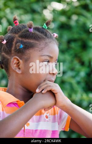 Portrait eines glücklichen kleinen afrikanischen Mädchens mit mehreren bunten Pferdeschwanzen, die im Garten in ihrem Haus in der Township stehen Stockfoto