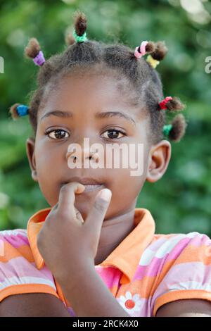 Portrait eines glücklichen kleinen afrikanischen Mädchens mit mehreren bunten Pferdeschwanzen, die im Garten in ihrem Haus in der Township stehen Stockfoto