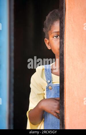 Schüchternes kleines afrikanisches Mädchen mit Zöpfen, die sich im Türrahmen in ihrem Haus in der Township verstecken Stockfoto