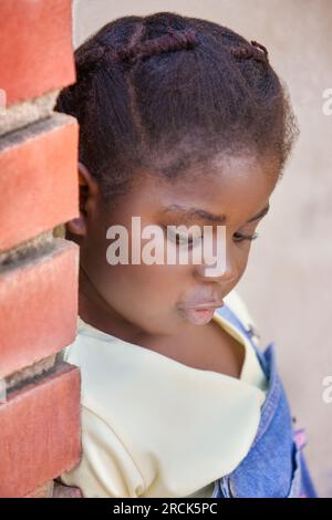 Kleines afrikanisches Mädchen mit Zöpfen, die sich an eine Backsteinwand lehnt, in ihrem Haus in der Township Stockfoto