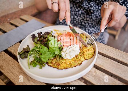 Frau schneidet Zucchini-Pfannkuchen mit Avocado, Käsesahne, Lachs und Ei. Gesundes Frühstück, Eiweiß. Restaurant-Gericht. Frühstück im Café bei Sonnenschein Stockfoto