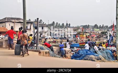 Ein Foto von Gebäuden und einer Gruppe junger Männer, die die Fischerboote und die Männer beobachten, die an Bord in Kapküste, Ghana, Westafrika arbeiten. Stockfoto