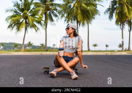 Junge, schöne Frau mit Mütze, die auf einem Skateboard sitzt und Kaffee trinkt Stockfoto