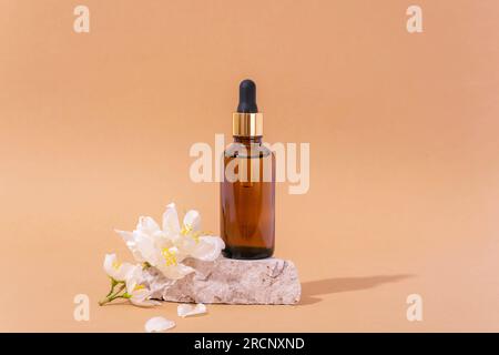 Braune Kosmetikserumflasche auf Steinpodium und weiße Jasminblumen auf neutralem beigefarbenem Hintergrund. Nahaufnahme. Stockfoto