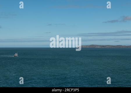 Einsames Fischerboot auf einem ruhigen blauen Meer mit fernem Land am Horizont unter klarem Himmel, Irische See, Großbritannien Stockfoto