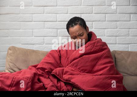 Kranke junge afrikanische Frau, die sich mit einer Decke bedeckt fühlt, sitzt auf dem Sofa, krankes schwarzes Mädchen, das zu Hause friert und sich erwärmt, eingewickelt mit Karo, keine Zentr Stockfoto