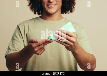 Glücklicher junger Mann, der sich eine Gesichtsmaskencreme auf die Finger legt, bevor er sie während seiner Hautpflege auf sein Gesicht aufträgt. Ein Mann, der sich mit einem um seine Haut kümmert Stockfoto