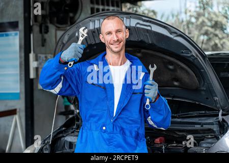 Ein glücklicher männlicher Mechaniker hält eine Ranch in jeder Hand, um zu feiern, während er mit offener Motorhaube vor einem Auto steht Stockfoto