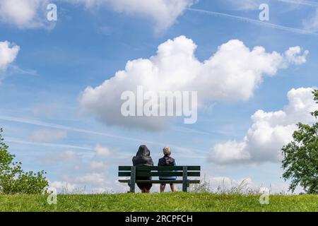 Blick aus dem niedrigen Winkel auf zwei Personen, die auf einer Parkbank auf einem Deich in den Niederlanden sitzen und in unsichtbarer Entfernung mit großen Cumuluswolken darüber blicken Stockfoto