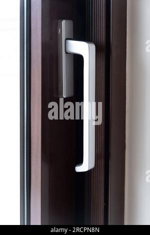 Silberner Aluminiumgriff an der geschlossenen Tür, Fenster vor braunem Rahmenhintergrund. Nahaufnahme der modernen, stilvollen und sicheren Struktur. Vertikal Stockfoto