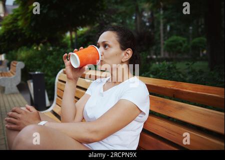 Junge, multiethnische Frau in weißem T-Shirt, Tourist, die Kaffee zum Mitnehmen aus einem umweltfreundlichen Einweg-Becher aus Papptasse trinkt, sitzt auf einem Holzmantel Stockfoto