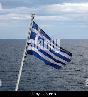 Griechenland steht für die führende Schifffahrtsmacht der Welt. Die griechische Flagge auf dem Fahnenmast winkte über dem gezackten Meerwasser. Wolkiger Himmelshintergrund. Stockfoto