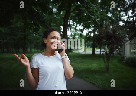 Wunderschöne, multiethnische junge Frau in einem weißen T-Shirt, lächelnd, telefonisch, während sie an einem Sommertag durch die Gasse eines Stadtparks schlendert. Menschen Stockfoto