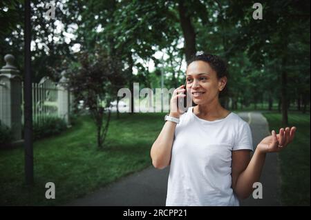 Attraktive junge afroamerikanische Frau in weißem T-Shirt, lächelnd, telefoniert am Sommertag auf der Gasse des Stadtparks. Peo Stockfoto