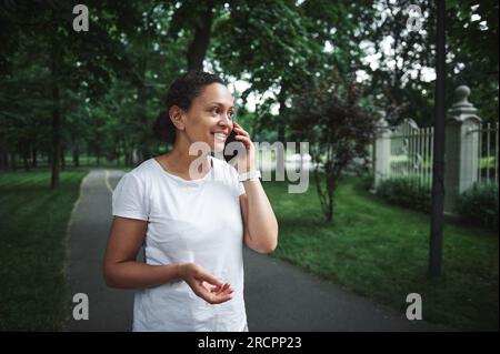 Wunderschöne, multiethnische junge Frau in einem weißen T-Shirt, lächelnd, telefonisch auf dem Smartphone, während sie an einem Sommertag durch die Gasse eines Stadtparks schlendert. Menschen. Stockfoto